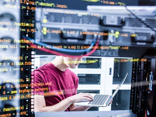 Person in the data centre working with a laptop between server cabinets, overlaid with programme code on the screen.