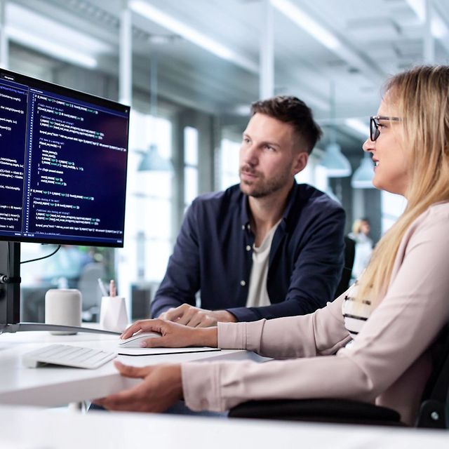 Two people are looking at source code on large monitors in a modern office and discussing it.