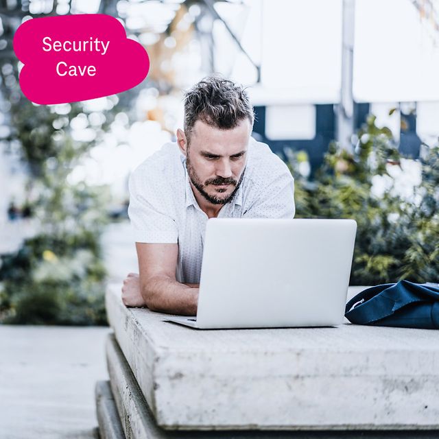 Person working outdoors lying down with a laptop on a concrete edge, green plants in the background.
