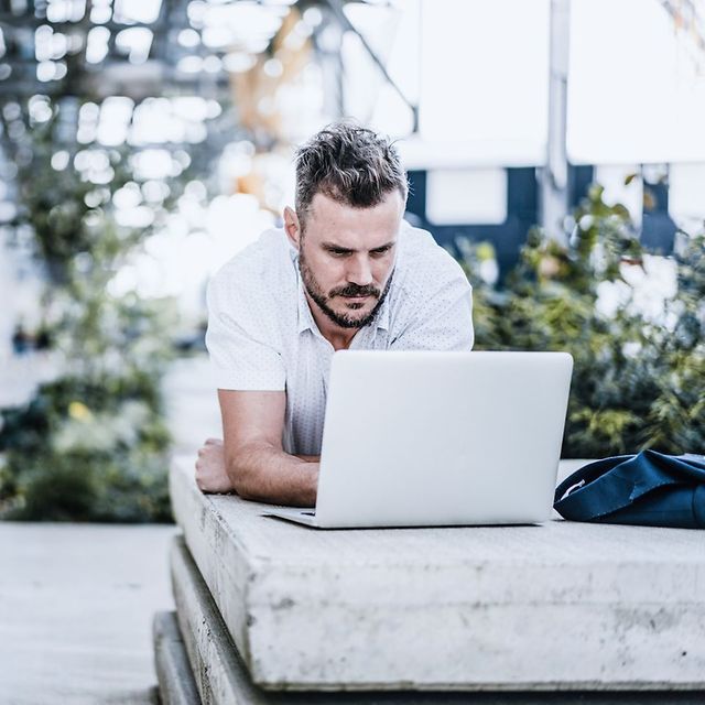 Person working outdoors lying down with a laptop on a concrete edge, green plants in the background.