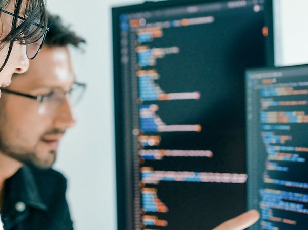 Two people analyse source code on large monitors, one points at the screen.