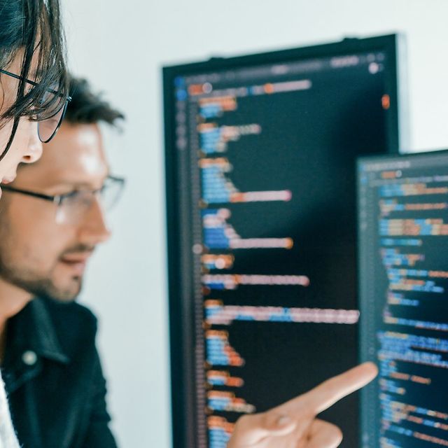Two people analyse source code on large monitors, one points at the screen.