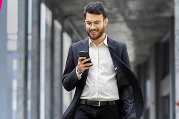 A young man in a white shirt and an open black jacket is walking with a mobile phone in his hand