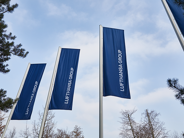 Three blue flags bearing the white lettering ‘Lufthansa’ are fluttering in the wind on flagpoles against a blue sky