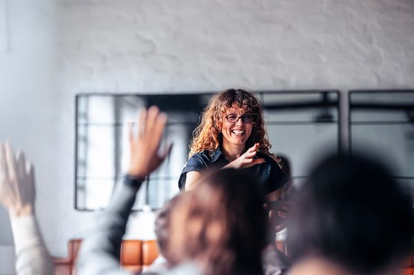 Frau moderiert ein Meeting, während Teilnehmende die Hand heben. Die Szene wirkt offen, aktiv und kollaborativ.