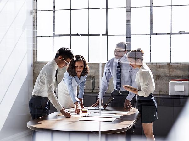 A working group in a modern office is gathered around a table discussing documents; photographed through a glass wall