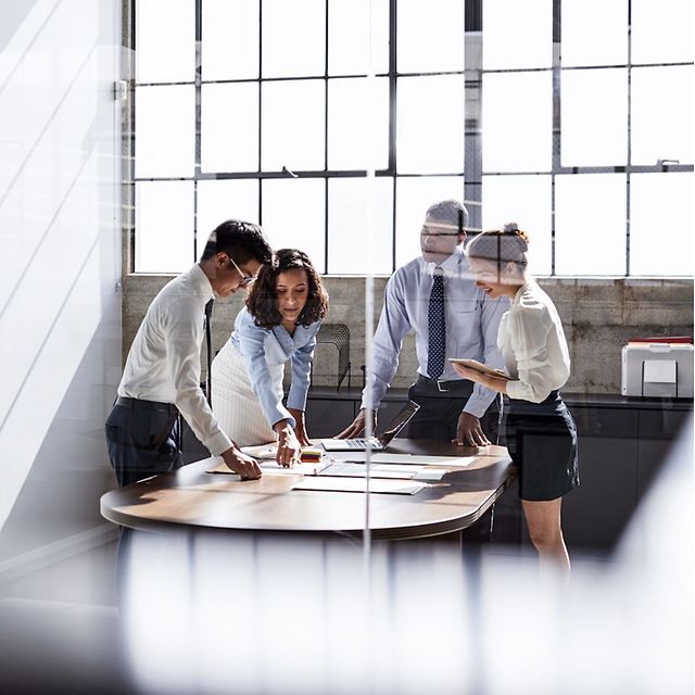 A working group in a modern office is gathered around a table discussing documents; photographed through a glass wall