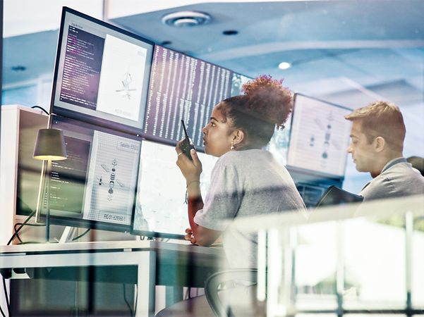 Two IT specialists are analysing data on several monitors in a control room.