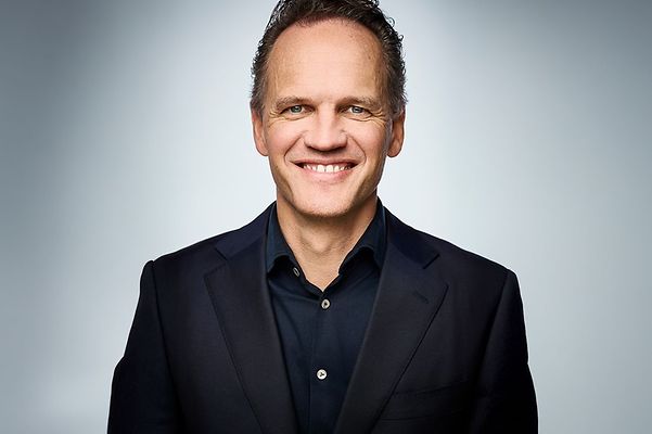 Portraitfoto von Thought Leader T Cloud Herr Lars Neumann lächelnd in schwarzem Hemd und Blaser vor grauem Hintergrund