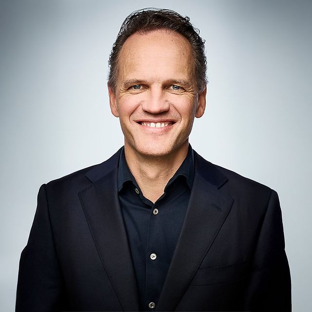 Portraitfoto von Thought Leader T Cloud Herr Lars Neumann lächelnd in schwarzem Hemd und Blaser vor grauem Hintergrund