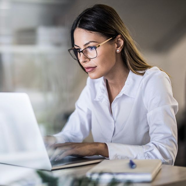 Frau mit Brille und weißer Bluse arbeitet konzentriert am Laptop im Büro, Notizbuch und Stift liegen daneben.