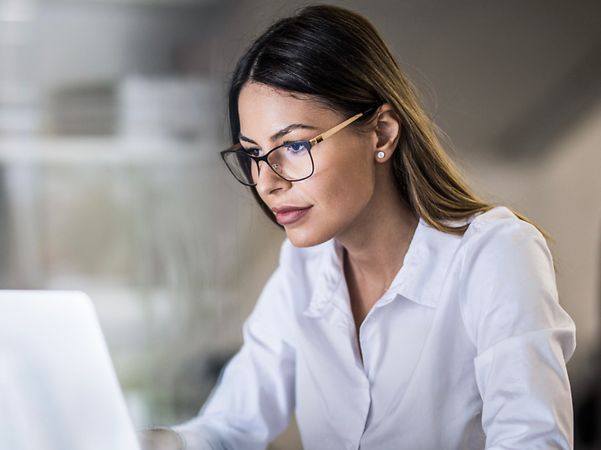 A woman wearing glasses and a white blouse is working intently on her laptop in the office; a notebook and pen lie beside her.