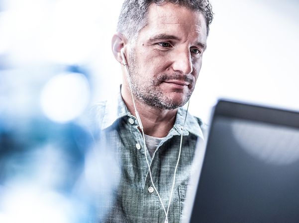 Mann mit Kopfhörern tippt konzentriert am Laptop, davor unscharfe blaue Lichtreflexionen (Bokeh).