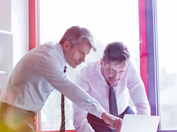 Zwei Geschäftsmänner in Hemd und Krawatte blicken auf einen Laptop, einer zeigt auf den Bildschirm, helles Büro