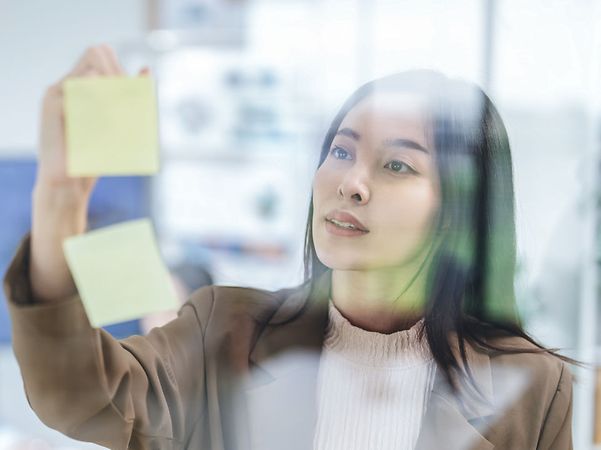Frau im Business-Blazer klebt gelbe Haftnotizen an eine Glaswand im Büro.