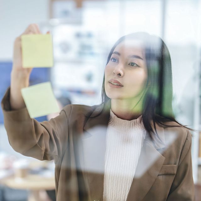 A woman in a business blazer is sticking yellow sticky notes to a glass wall in the office.