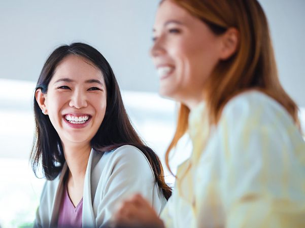 Zwei lachende Frauen sitzen nebeneinander an einem Tisch in einem hellen Büro, im Vordergrund ein Laptop unscharf.