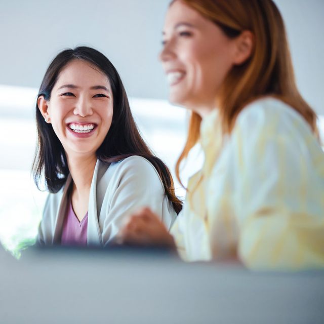 Zwei lachende Frauen sitzen nebeneinander an einem Tisch in einem hellen Büro, im Vordergrund ein Laptop unscharf.