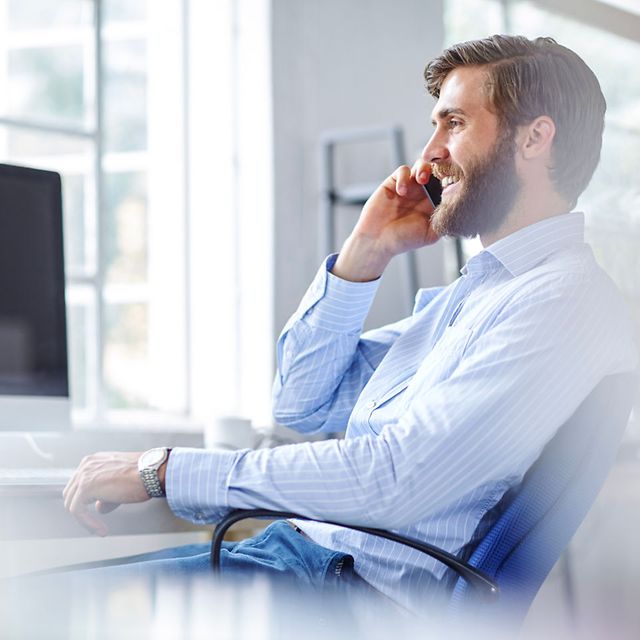 Lächelnder Mann im blauen Hemd telefoniert mit dem Smartphone am Schreibtisch in einem hellen Büro.