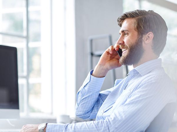 A smiling man in a blue shirt is on the phone with his smartphone whilst sitting at a desk in a bright office.