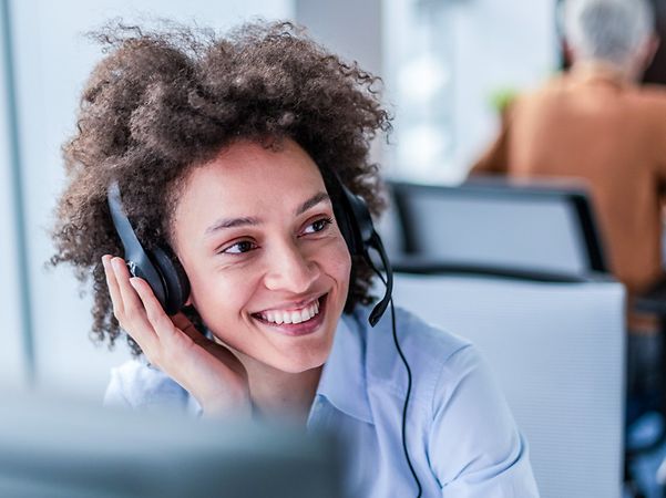 Lächelnde Frau mit Headset und lockigem Haar im Büro, blickt zur Seite; im Hintergrund arbeitet ein Kollege am PC.