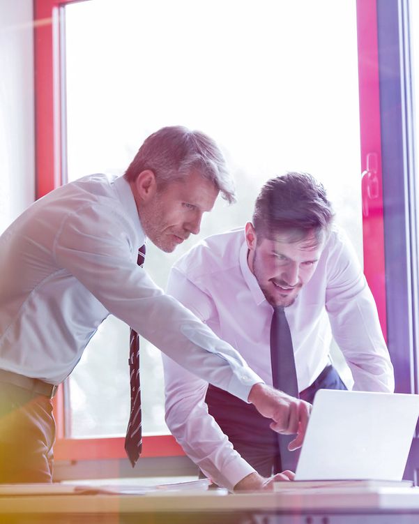 Two businessmen in shirts and ties are looking at a laptop; one is pointing at the screen; a brightly lit office