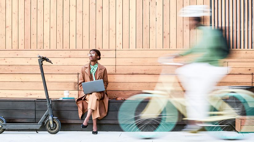 Frau sitzt mit Notebook auf einer Bank, ein Radfahrer fährt im Vordergrund durchs Bild