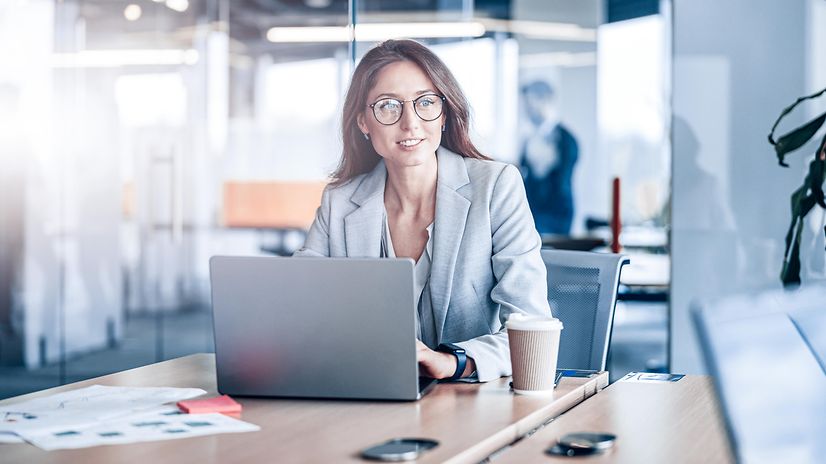 Lächelnde Geschäftsfrau mit Brille, die in einem modernen Büro in einem Unternehmen an einem Laptop arbeitet