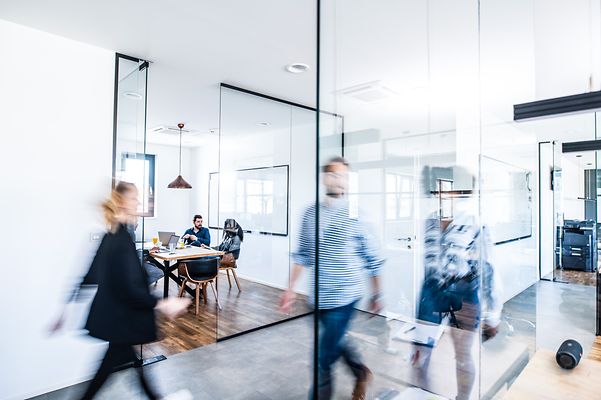 Kollegen sitzen im Meetingraum, im Vordergrund gehen andere Kollegen als dynamisch verwischte Silhouetten durchs Bild