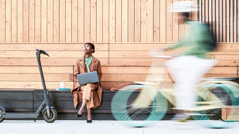 Eine Frau sitzt auf einer Bank und schaut ins Freie, ein Mann fährt Fahrrad.