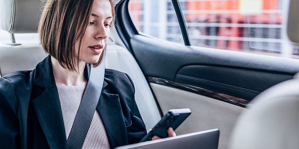 Frau sitzt mit ihrem Laptop und Smartphone im Auto und schaut auf den Bildschirm.