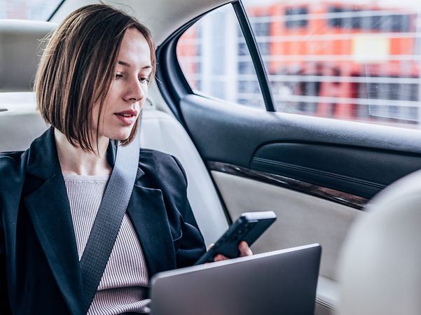 Frau sitzt mit ihrem Laptop und Smartphone im Auto und schaut auf den Bildschirm.