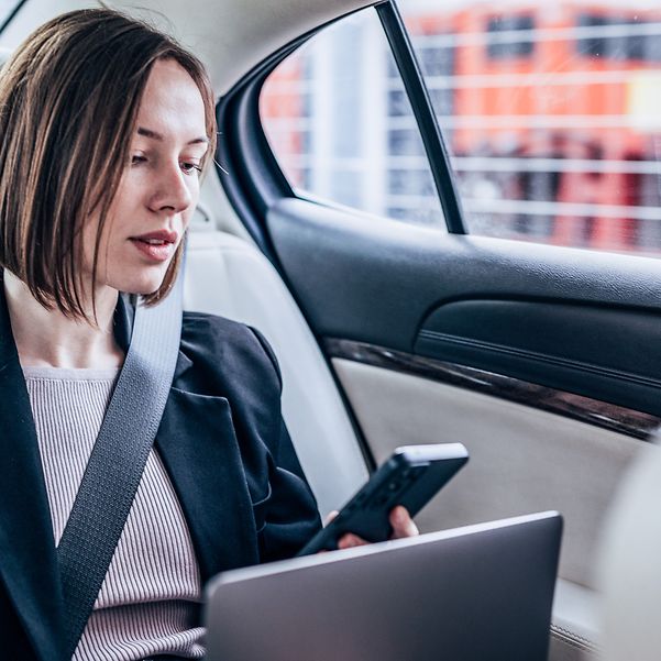 Frau sitzt mit ihrem Laptop und Smartphone im Auto und schaut auf den Bildschirm.
