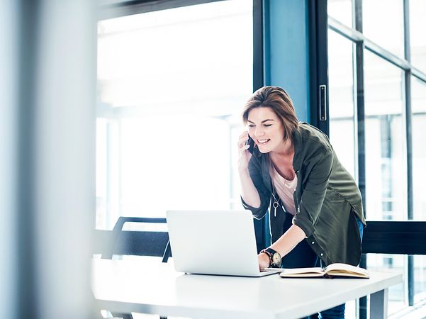 Eine Frau telefoniert in einem hellen Büro vor einem Laptop und kümmert sich um App-Modernisierung durch Hybrid-IT-Umgebungen
