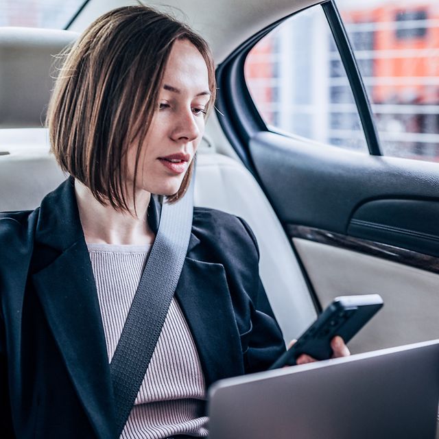 Business Analytics Frau sitzt in Ihrem Auto hinten und arbeitet an Ihrem Laptop und schaut auf Ihr Smartphone