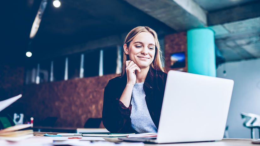 Eine Frau sitzt in einem modernen Büro an einem Tisch mit Unterlagen und schaut auf den Bildschirm.