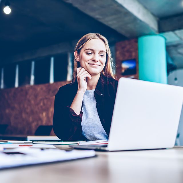 Eine Frau sitzt in einem modernen Büro an einem Tisch mit Unterlagen und schaut auf den Bildschirm.