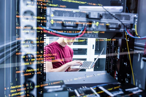 Ein junger Mann, der einen Laptop hält, steht im Serverraum und arbeitet an einem Server