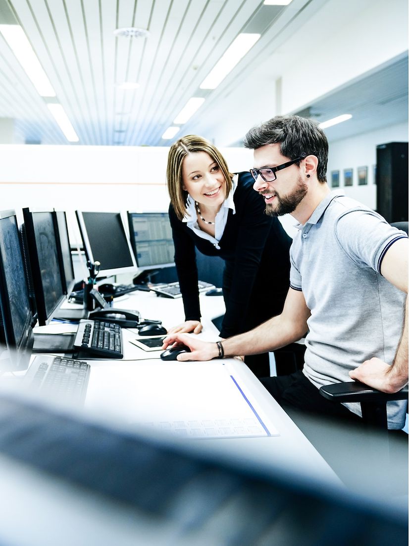 Zwei Personen, eine Frau und ein Mann, sprechen miteinander an einem Schreibtisch mit vielen Monitoren